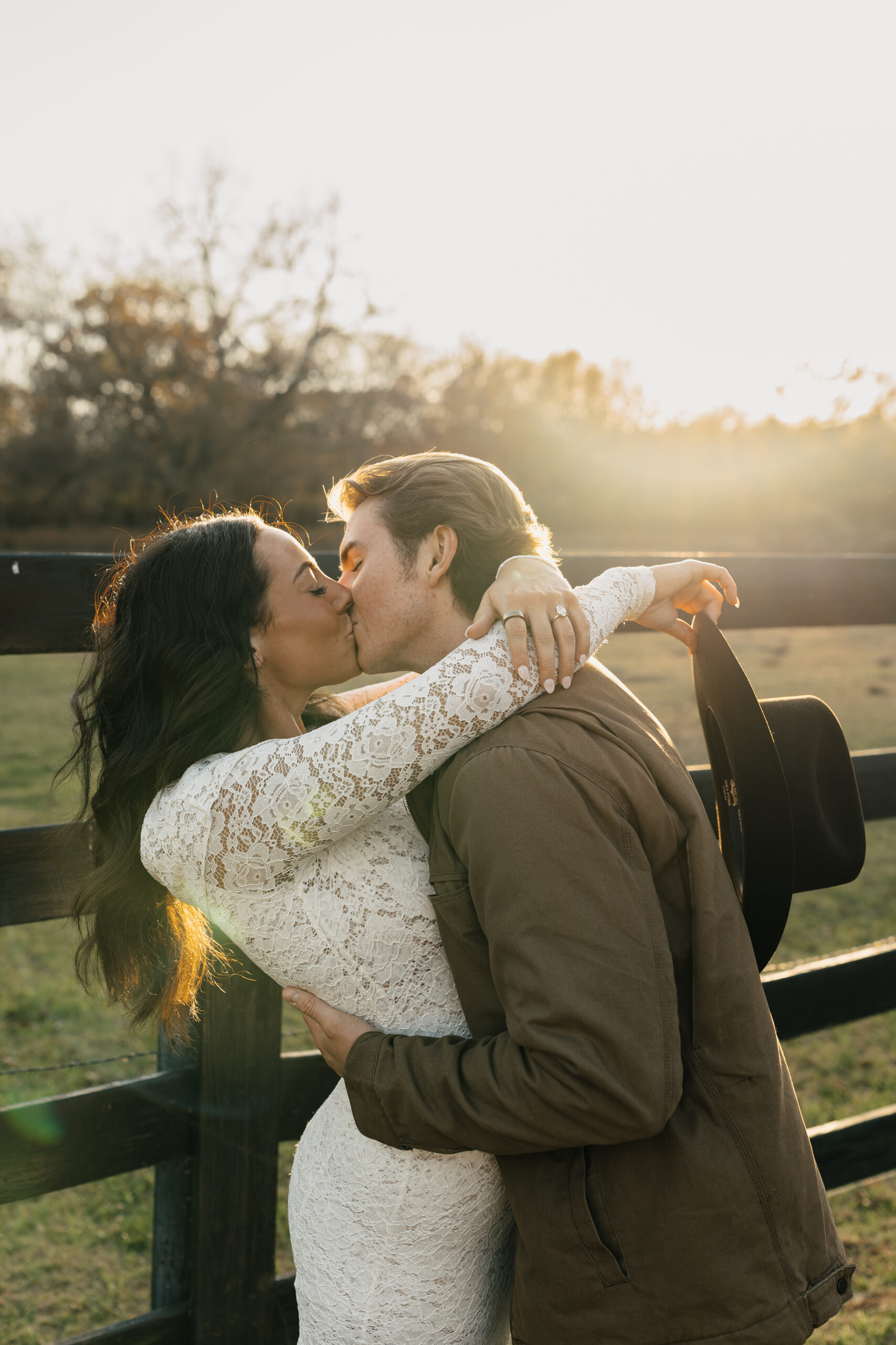 Fiancé leaning against a fence wall while bride leans on him, sun flares in the background